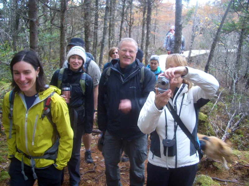 A small group of hikers stopped among bare hardwoods on a November walk, one of them holding a camera.